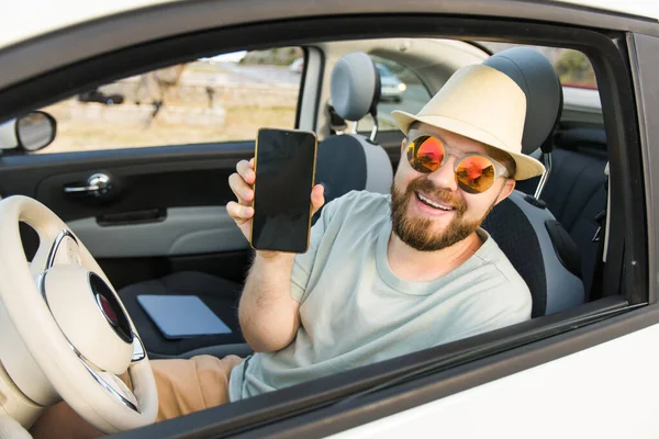 Smiling bearded man in sunglasses showing mobile phone with empty screen for mock up sitting on driver seat in new luxury car cabriolet, holding gadget in hand selective focus on device