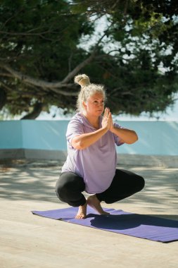 Sabahları deniz kenarında kıdemli bir kadın esneme pozisyonu alıyor. Yaşlı kadın plajda yoga yapıyor..