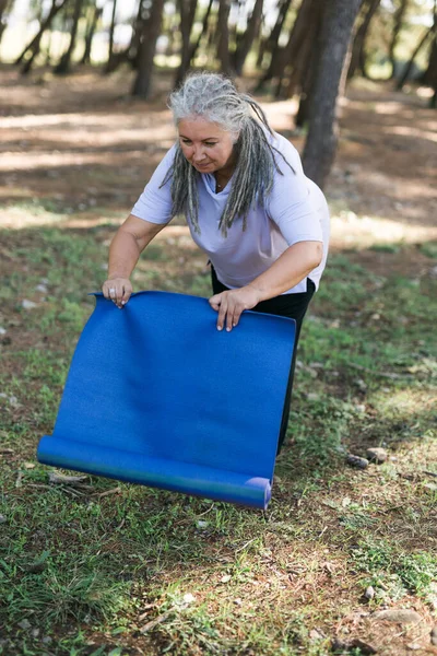 Old woman holding yoga mat and preparing to practice yoga or meditation outdoors on sea beach. Happy mature woman with dreadlocks exercising on seashore. Meditation or yoga and relaxation c