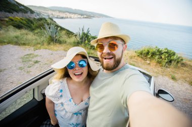 Mutlu ve güzel bir çift tatilde üstü açık bir arabayla selfie çektiriyorlar. Kiralık arabalar ve tatil konsepti