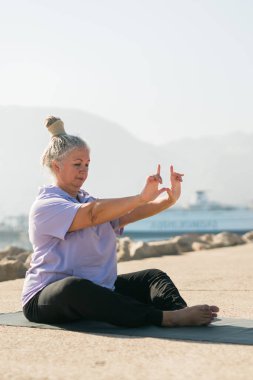 Ciddi bir saç stili olan yaşlı kadın güneşli bir günde poz verirken kollarını kaldırarak plajda dikiliyor.