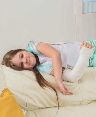 Kırık bileği ya da kolu olan bir çocuk gülümsüyor ve kanepede eğleniyor. Pozitif tutum, iyileşme ve çocuk.