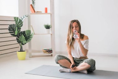 Matta yoga yapan genç ve güzel bir kız. Mola sırasında su içiyor. Fitness ve sağlıklı yaşam tarzı konsepti