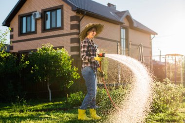 Beyaz kadın bahçıvan iş kıyafetleri içinde güneşli yaz gününde sebze bahçesindeki yatakları suluyor. Bahçede çalışma kavramı