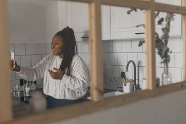 Happy modern African American young girl talking on video chat and sit home communication concept. Smiling hipster girl making self portrait on her telephone at home