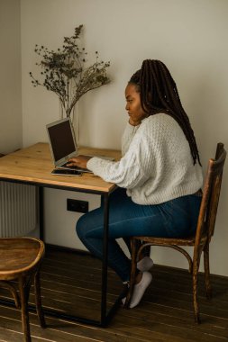 Cheerful african american woman using laptop while sitting on chair in living room - student, video call and working at home