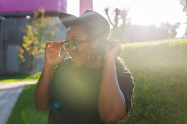 Tişört giyen mutlu Afrikalı Amerikalı kadın sokakta kameraya bakıp gülüyor. Z kuşağı hipster kızı güneş ışığı portresiyle arka planda poz veriyor.