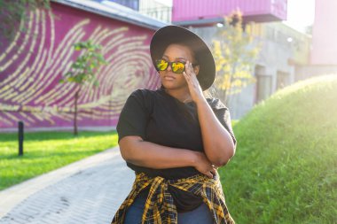 Tişört giyen mutlu Afrikalı Amerikalı kadın sokakta kameraya bakıp gülüyor. Z kuşağı hipster kızı güneş ışığı portresiyle arka planda poz veriyor.