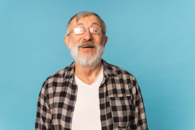 Beyaz saçlı, sakallı, ağzı açık emekli yaşlı bir adam mavi arka planda heyecanla gülüyor.