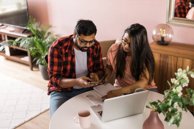 Serious wife and husband planning budget, checking finances, focused young woman using online calculator, counting bills or taxes, man using laptop, online banking services. Family sitting at table in