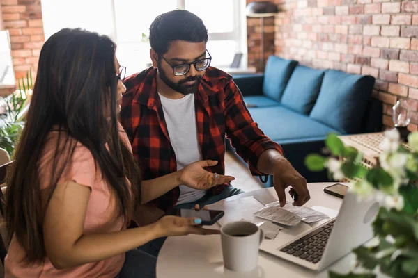 Young millennial indian husband and wife doing domestic paperwork, accounting job and reviewing paper bills, receipts at laptop computer, using online calculator and paying mortgage rent fees on