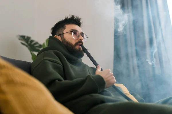 stock image Bearded millennial or gen z man smoking hookah while relaxing on sofa at home - chill time and resting