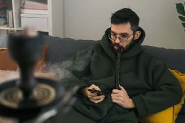 Bearded man is smoking hookah at home and blowing cloud of smoke - chill time and resting