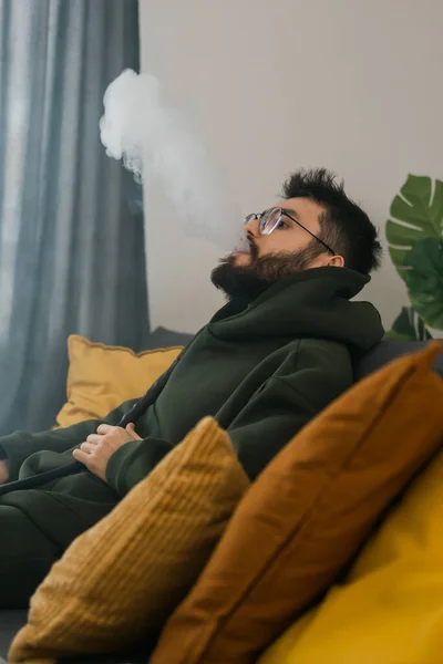 ひげを生やしたミレニアルまたはジェンZ男喫煙フカながらリラックスしている間にソファに自宅で クール時間と休息 — ストック写真