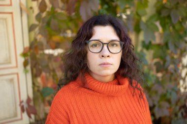 Turuncu kazak ve gözlük takan şık, mutlu genç bir kadın. Şehrin arka planında gözlüklü gülümseyen kız portresi