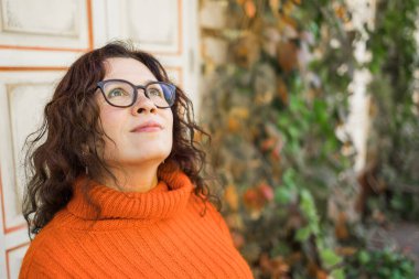 Autumn portrait of an attractive young woman in stylish glasses in a knitted fashionable orange sweater on background leaves. Girl walks in city
