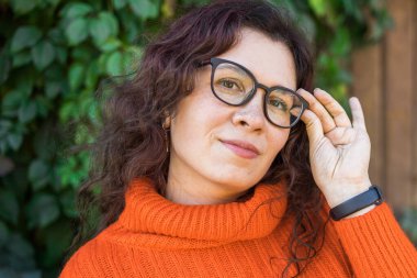 Autumn portrait of an attractive young woman in stylish glasses in a knitted fashionable orange sweater on background leaves. Girl walks in city