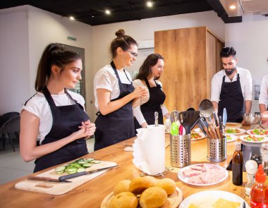 Aşçılık kursu konsepti - aşçı üniforması giyen kıdemli erkek aşçı, gençlere aşçılık sınıfı öğrencilerine yemek pişirmeyi ve mutfak malzemelerini karıştırmayı öğretiyor.