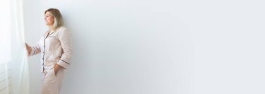 Gülümseyen sarışın kadın pijamayla oturma odasında rahat bir koç üzerinde oturuyor. Ev kıyafetleri ve evde boş zaman tatili.