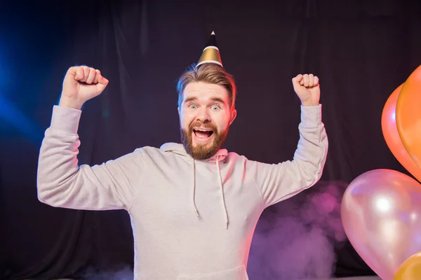 Joyful birthday man wears paper cone hat dancing and sing and stands near colorful helium balloons enjoys party and expresses happiness - holidays and winner