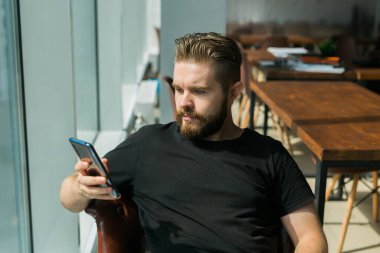 Gülümseyen genç serbest çalışan iş adamı cep telefonu kullanıyor sosyal medya ağlarını kontrol ediyor ya da sandalyede oturan mesajları dinliyor. Çevrimiçi dijital iletişim ve aygıt bağımlılığı