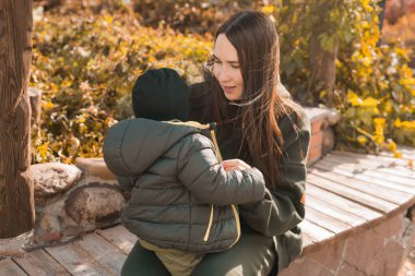 Anne ve küçük oğlu sonbahar parkında. Kadın çocuk ceketinin fermuarını çeker.