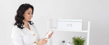 Beyaz elbiseli genç Hintli kadın beyaz arka planda kozmetik ürünü şişesini tutarken gülümsüyor..