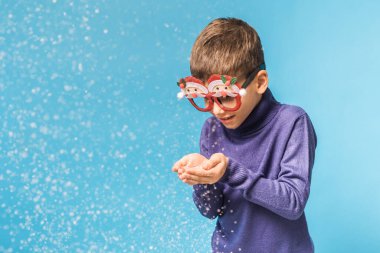 Çocuk mavi arka planda ellerinden kar üflüyor. Noel ve yeni yıl tatili konsepti. Nesil alfa ve gen alfa çocukları