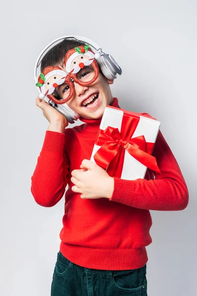 Child boy holds xmas gift box listening to Christmas music with funny Santa Claus glasses on white