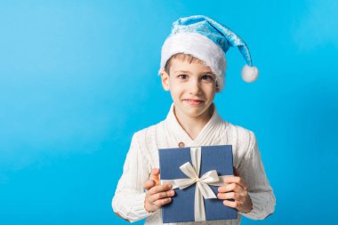 Hediye kutusu fotokopi alanı olan mavi Noel şapkalı küçük mutlu çocuk. Mesajın için boş yer. Kış tatili ve şimdiki konsept