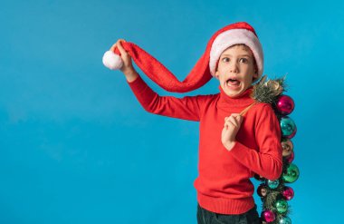 Noel Baba şapkalı neşeli çocuk mavi arka planda Noel süslemeleri, Noel ve tatil konsepti modellemeleri yapıyor. Nesil alfa ve gen alfa çocukları