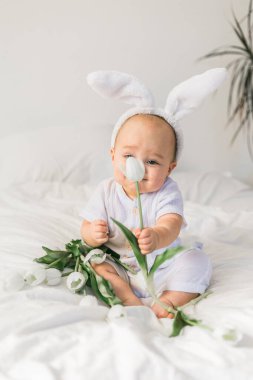 Tavşan kulaklı, evde beyaz bir yatak üzerinde laleleri olan sevimli, komik bir bebek. Paskalya tebrik kartı.