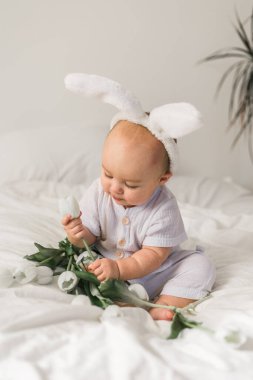 Tavşan kulaklı, evde beyaz bir yatak üzerinde laleleri olan sevimli, komik bir bebek. Paskalya tebrik kartı.