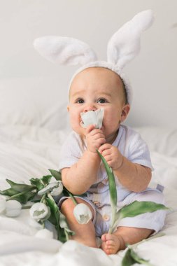 Tavşan kulaklı, evde beyaz bir yatak üzerinde laleleri olan sevimli, komik bir bebek. Paskalya tebrik kartı.