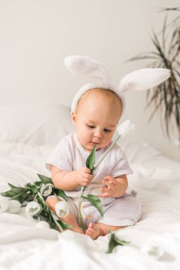 Tavşan kulaklı, evde beyaz bir yatak üzerinde laleleri olan sevimli, komik bir bebek. Paskalya tebrik kartı.