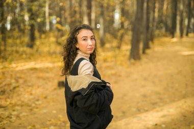 Sonbahar parkında yürüyen çekici genç bir kadın, mutlu bir ruh hali ve moda tarzı ve kıvırcık uzun kahverengi saçlar. Sonbahar sezonu ve güzel kadın portresi. Milenyum nesli