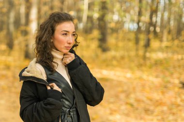 Sonbahar parkında yürüyen çekici genç bir kadın, mutlu bir ruh hali ve moda tarzı ve kıvırcık uzun kahverengi saçlar. Sonbahar sezonu ve güzel kadın portresi. Milenyum nesli