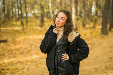 Sonbahar parkında yürüyen çekici genç bir kadın, mutlu bir ruh hali ve moda tarzı ve kıvırcık uzun kahverengi saçlar. Sonbahar sezonu ve güzel kadın portresi. Milenyum nesli