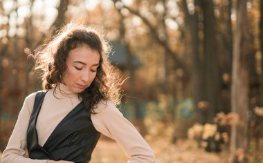 Sonbahar parkında yürüyen çekici genç bir kadın, mutlu bir ruh hali ve moda tarzı ve kıvırcık uzun kahverengi saçlar. Sonbahar sezonu ve güzel kadın portresi. Milenyum nesli