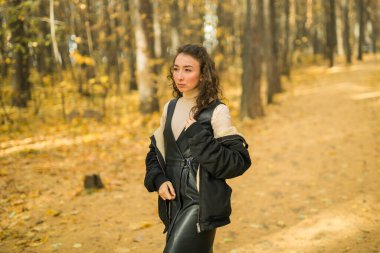 Sonbahar parkında yürüyen çekici genç bir kadın, mutlu bir ruh hali ve moda tarzı ve kıvırcık uzun kahverengi saçlar. Sonbahar sezonu ve güzel kadın portresi. Milenyum nesli