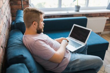 Milenyum adamı evdeki kanepede oturmuş internette dizüstü bilgisayarla çalışıyor ve internet kullanıyor. Gülümseyen ve mutlu ruh hali, serbest çalışan ve serbest zaman, rahat ve modern iş hayatı konsepti. Boşluğu kopyala