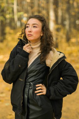 Sonbahar parkında yürüyen çekici genç bir kadın, mutlu bir ruh hali ve moda tarzı ve kıvırcık uzun kahverengi saçlar. Sonbahar sezonu ve güzel kadın portresi. Milenyum nesli