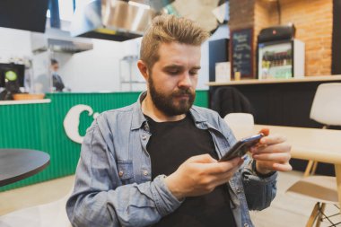 Bearded male using smartphone at modern coffee shop and chatting online messaging on mobile phone. Social networks and reading blog