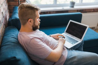 Milenyum adamı evdeki kanepede oturmuş internette dizüstü bilgisayarla çalışıyor ve internet kullanıyor. Gülümseyen ve mutlu ruh hali, serbest çalışan ve serbest zaman, rahat ve modern iş hayatı konsepti. Boşluğu kopyala