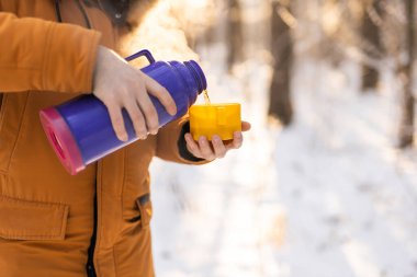 Man pours hot tea from a thermos into a snow walking in snowy frozen winter forest at sunset. Adventure, tourism and camping concept. Copy space and empty place for text advertising.