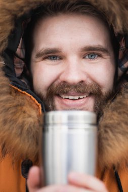 Close-up portrait handsome bearded millennial man in winter clothes and with thermos snow outdoor. Cold season and hot beverage in winter time.