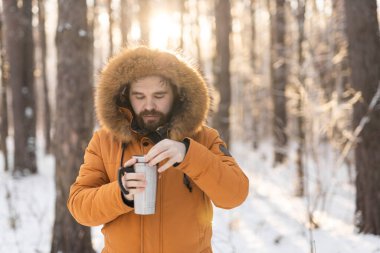 Close-up portrait handsome bearded millennial man in winter clothes and with thermos snow outdoor. Cold season and hot beverage in winter time.