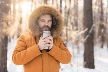 Close-up portrait handsome bearded millennial man in winter clothes and with thermos snow outdoor. Cold season and hot beverage in winter time.