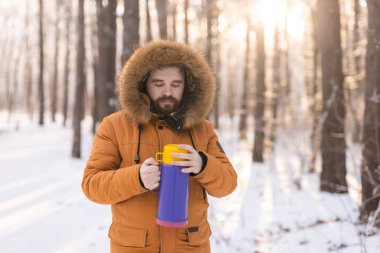 Close-up portrait handsome bearded millennial man in winter clothes and with thermos snow outdoor. Cold season and hot beverage in winter time.