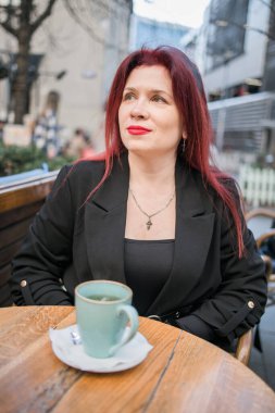 Restoranın terasında parlak kırmızı rujlu güzel bir kadın oturuyor. Kahvede poz veren siyah elbiseli kızıl saçlı harika bir kadın.